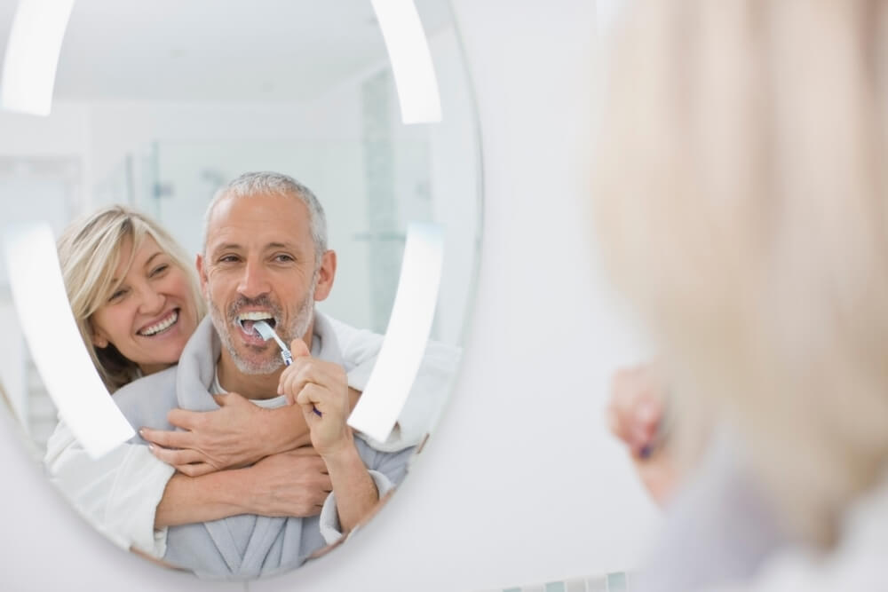 Older man brushing his teeth while older woman smiles and hugs him