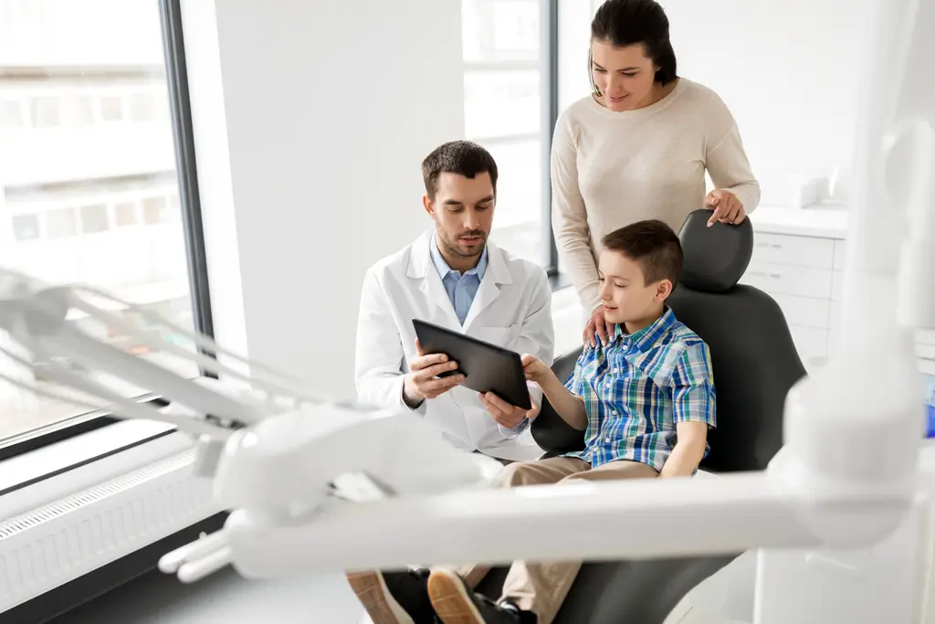 Male dentist showing a treatment plan on a tablet to a young boy and his mother in a modern dental office.
