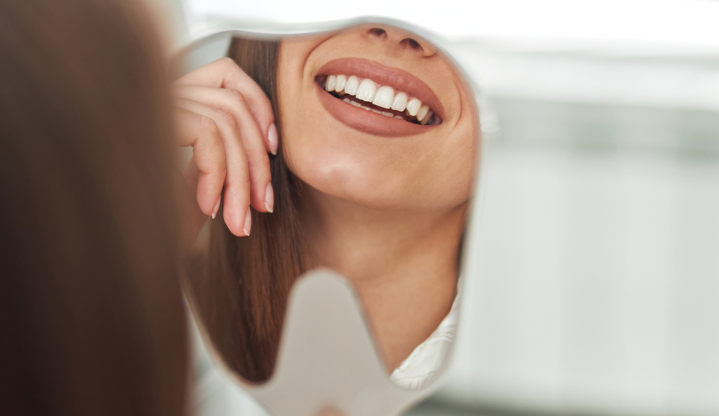 Close-up of a woman smiling at her reflection in a dental mirror, showing straight white teeth.